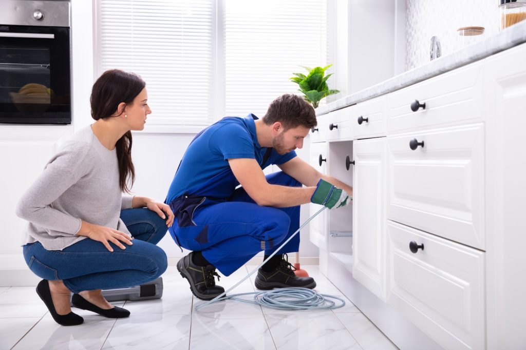 A homeowner standing by as a plumber performs drain cleaning in the kitchen sink.